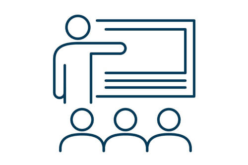 Meet our faculty. Icon depicts a person pointing to a chalkboard while standing in front of a class .