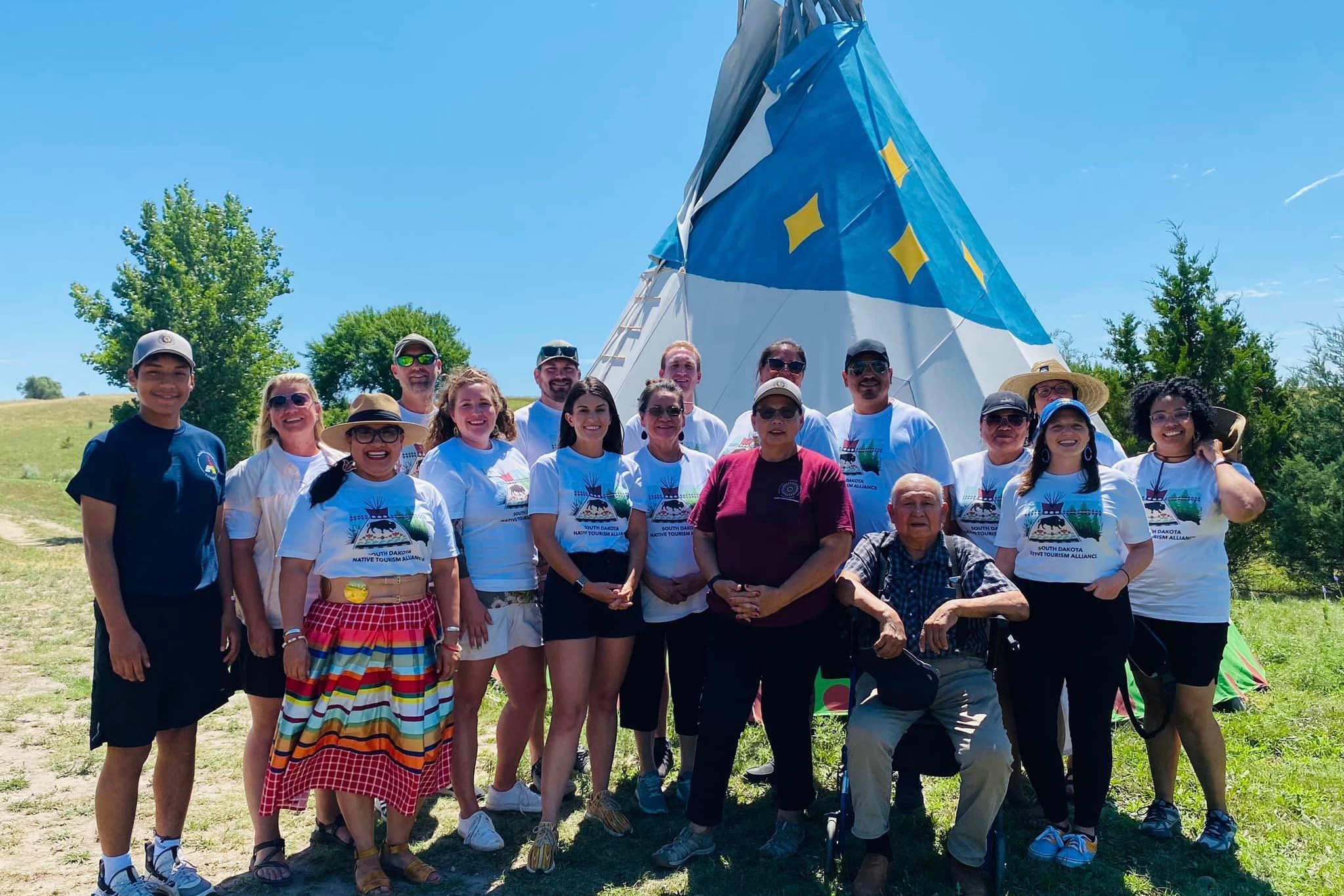 A group of approximately 20 people stand and pose for a photo outdoors. They wear matching shirts that read "South Dakota Native Tourism Alliance." The landscape is has grass and trees. A tepee can be seen behind the group.