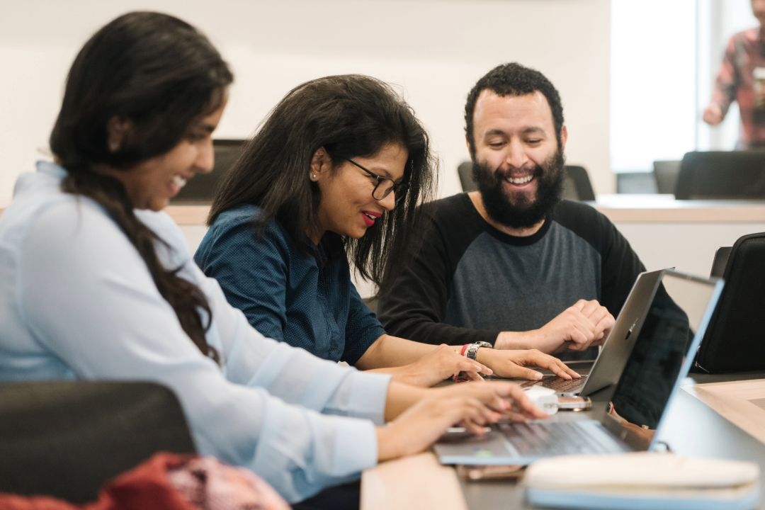 A group of three people sit and share a laugh. They look at laptops. 