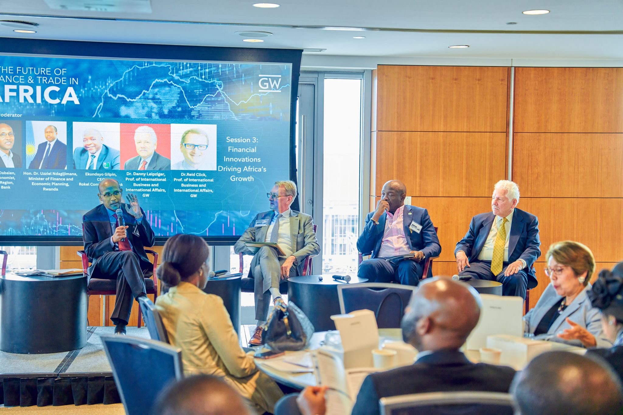 Four people sit on a stage. They wear business attire. One is speaking with a microphone . An audience watches them. A projector displays the word Africa and images of the speakers.