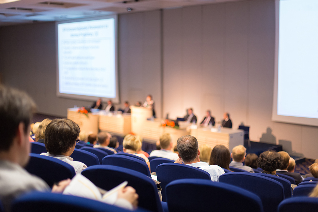 audience members in an auditorium look down from raised seats to a panel of speakers on stage below