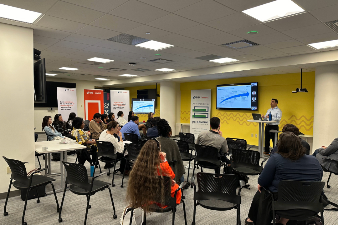 Students listen to a speaker at the front near a digital screen, inside a brightly lit room with banner displays.