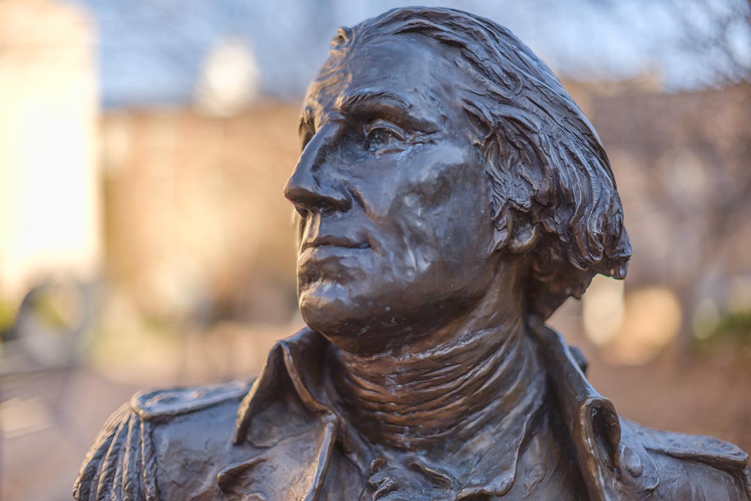 Image shows a broze statue of President George Washington with the GW University campus in the background.