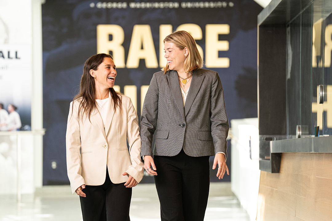 two smiling women walk and chat in front of a GW banner that reads "raise high"