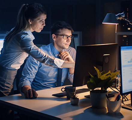 in a darkened office, a woman gestures toward a computer screen as a man looks on