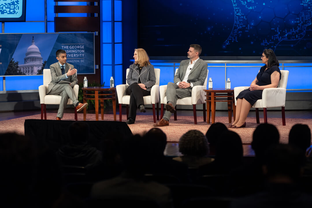 Four people seated in chairs on a stage indoors. An audience appears.