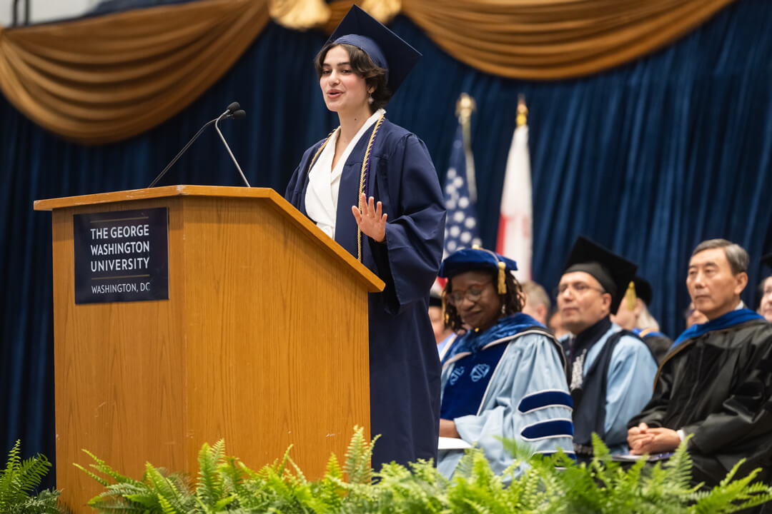 Anna Shah speaking in front of a podium with the George Washington University logo. She and those in the audience wear graduation regalia.