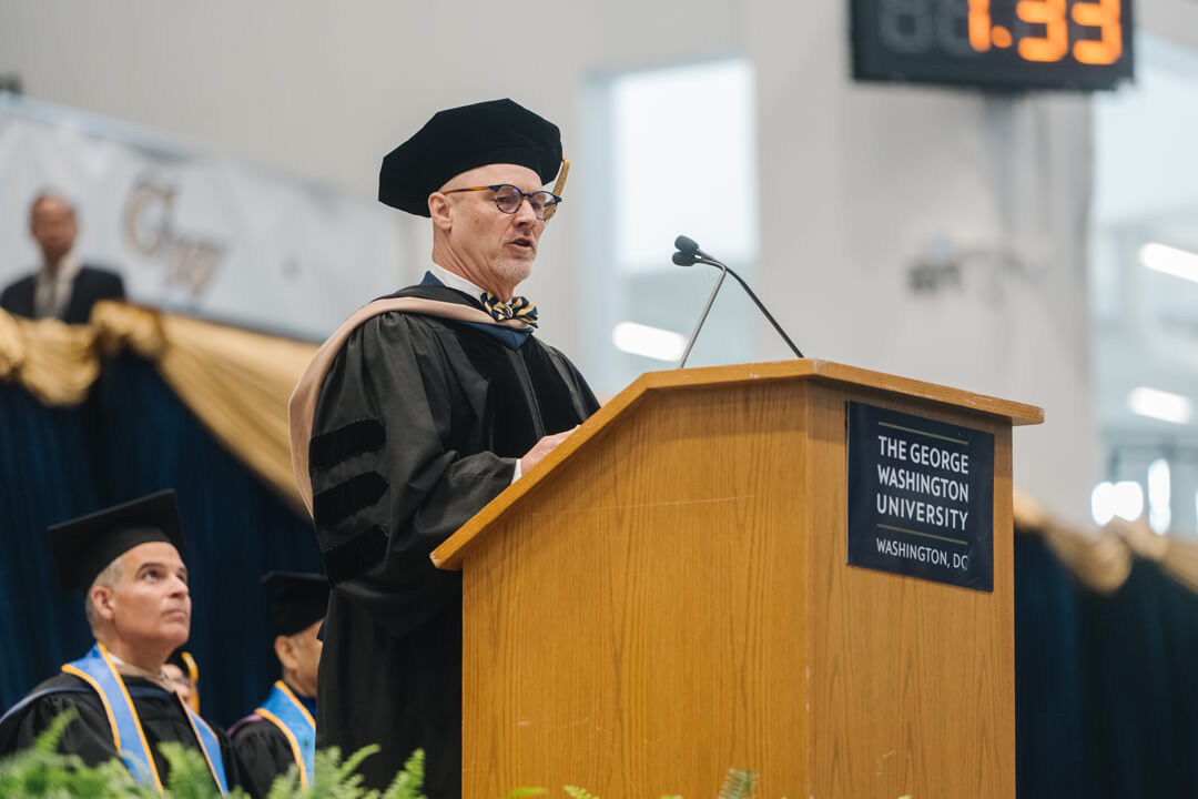 John Roberts, an alumnus of the GW School of Business speaks in front of a podium. He wears regalia.  Others can be seen  on the platform behind him. 