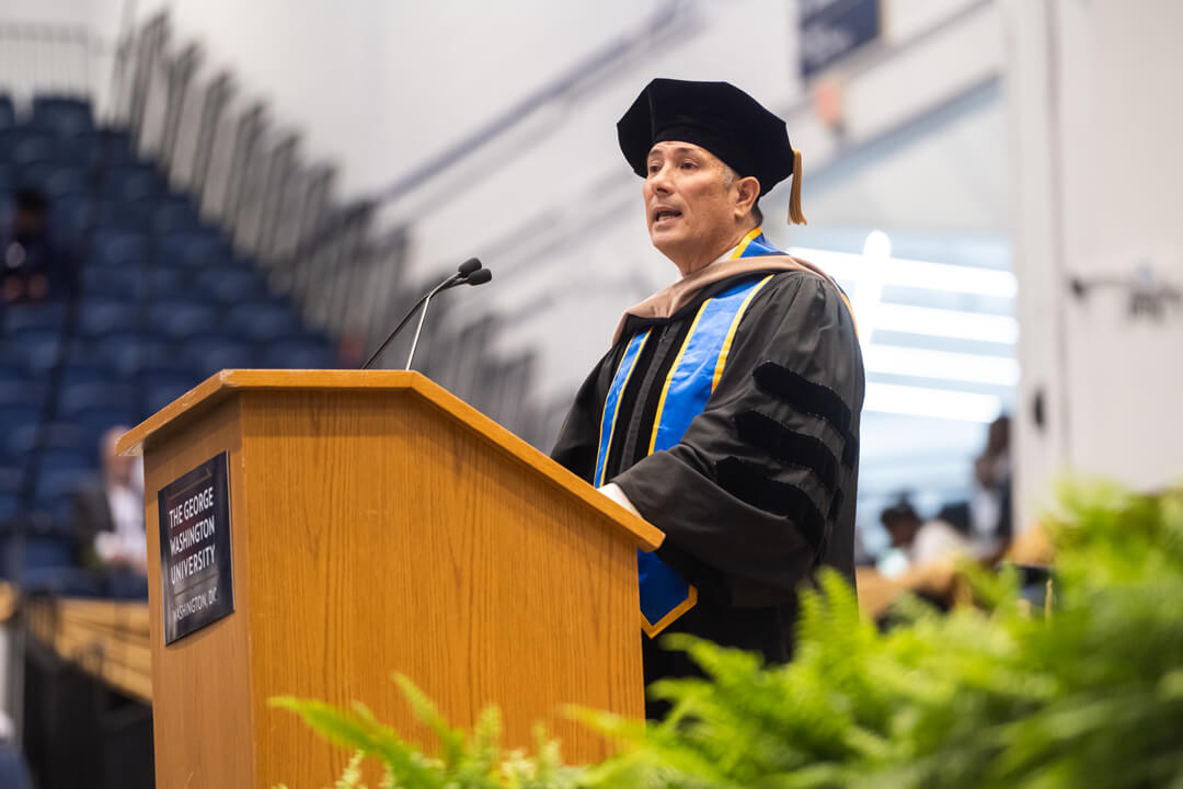 Greg Wong speaks in front of a podium with a microphone. He wears graduation regalia, including a cap and gown.  He appears indoors. Decorations can be seen. 
