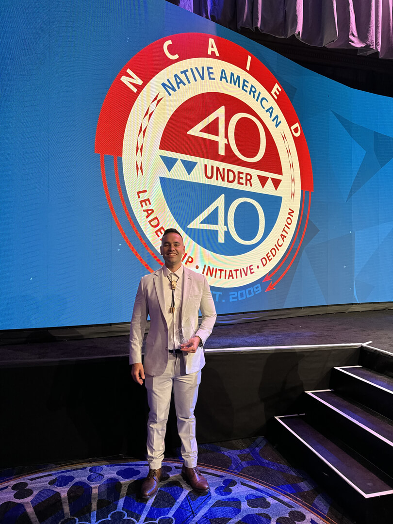  Quinton Uksi Carroll stands on a stage indoors. He wears a suite. A graphic with the words Native American 40 Under 40 appears behind him.