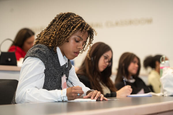 student writes in notebook during class, other students in the background