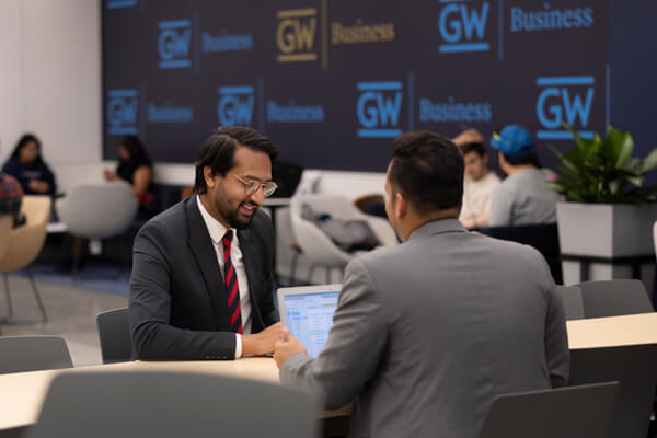 two students wearing suits talk across table, one has laptop open in front of him