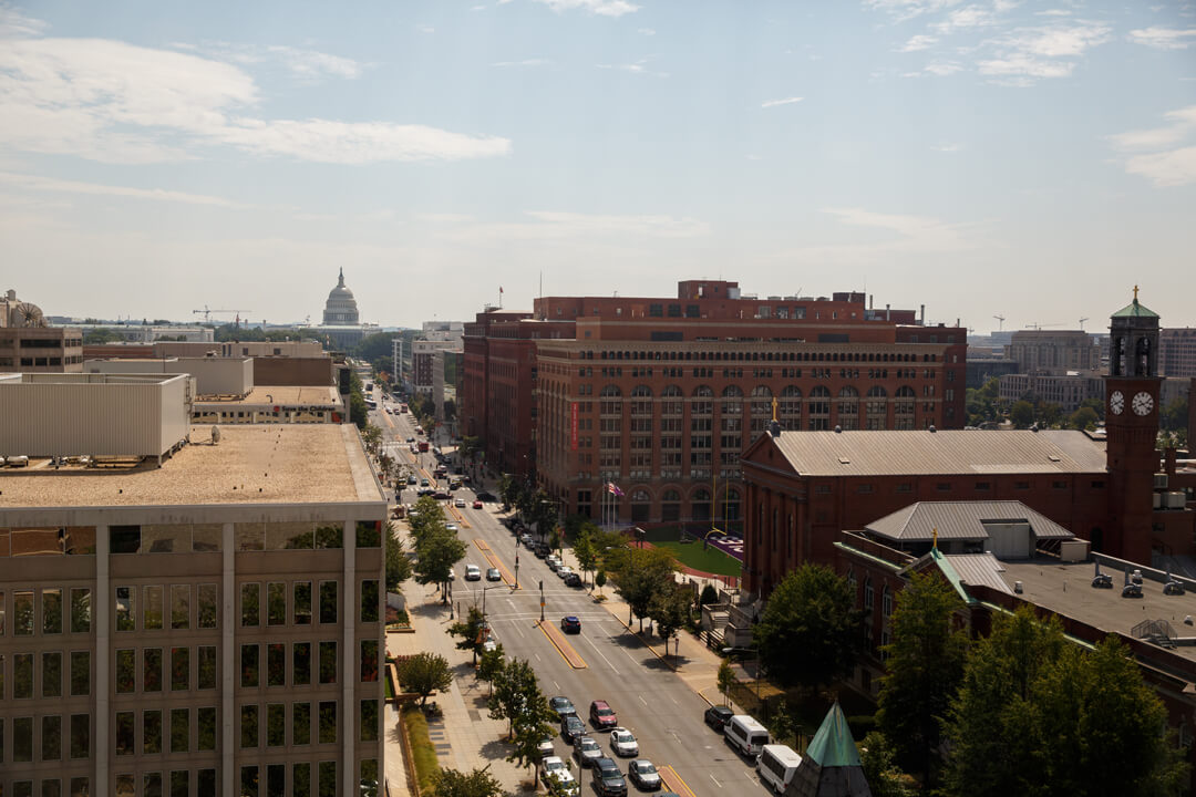 An aerial view of a busy Washington, D.C. street. A somewhat cloudy sky is visible. The U.S. Capitol building can be seen from afar.