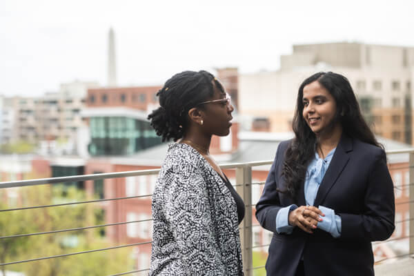 two students talk to each other on outdoor patio, campus buildings in the background