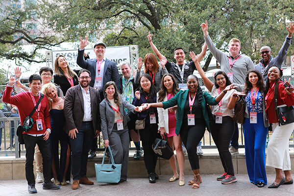 a large group of individuals pose for a group photo, all look excited, some have hands raised, tree in background