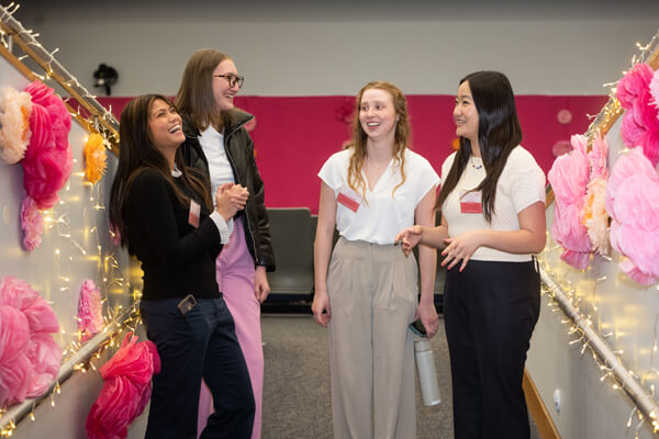 four business students laugh at event, pink paper flowers line the entry way to the sides of them