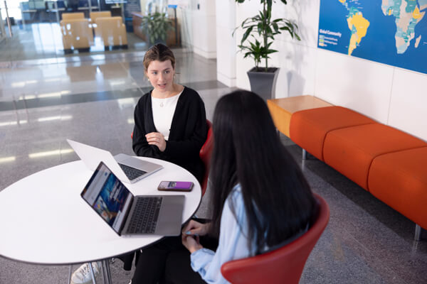 two GWSB students sit at a round table talking, laptops open in front of them