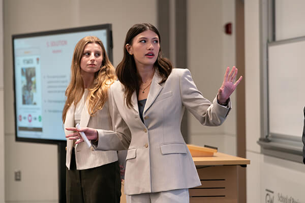 two female business students look and gesture toward presentation in front of class, screen out of camera view
