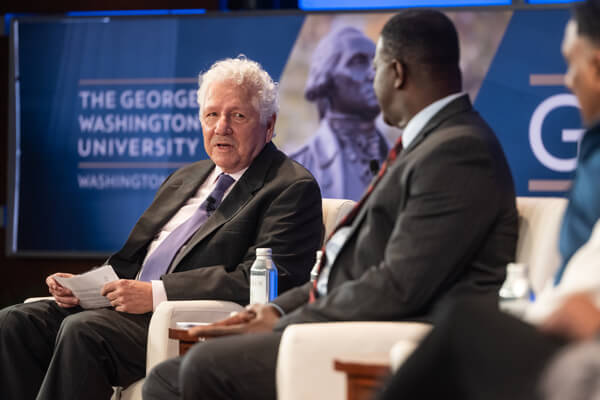 two panelists at event in discussion while seated in white chairs on stage