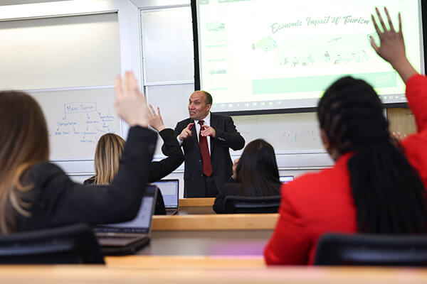 professor leads discussion in front of class, two students with backs to camera raise their hands