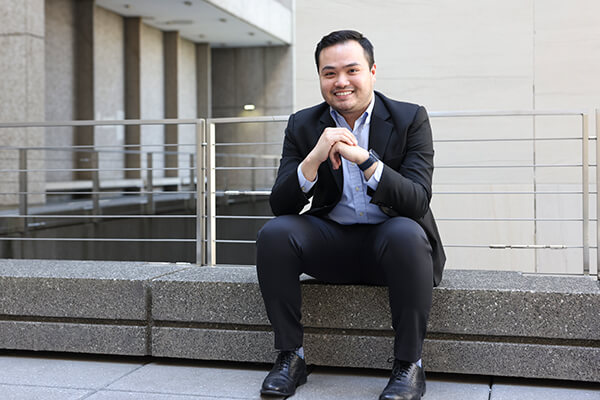 male wearing business suit sits on low concrete wall with railing outside business school
