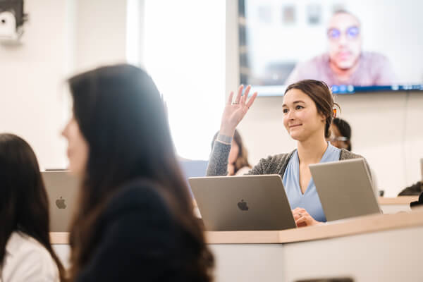 student in class raises her hand, laptop open in front of her