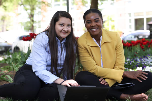 two professionally dressed students smile at the camera, sitting outside
