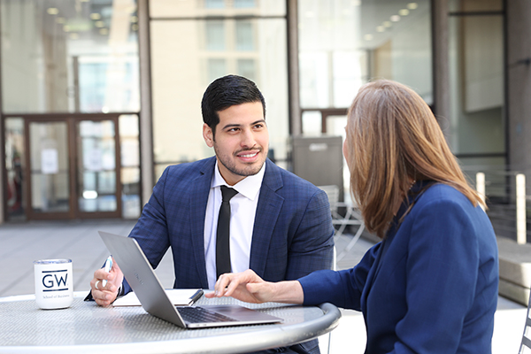two students sit to talk at outdoor table, laptop open in front of one, GW coffee cup next to another