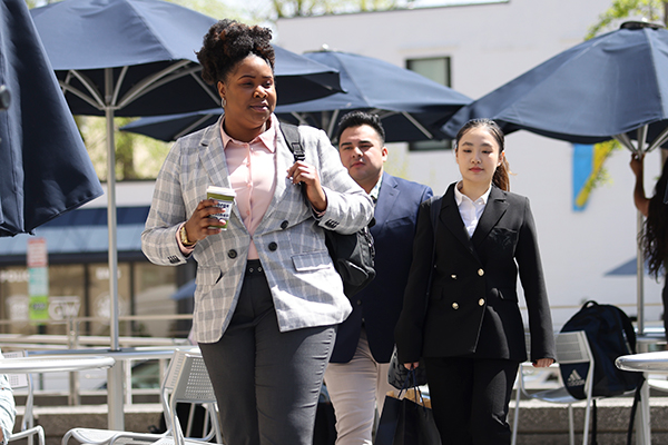 students walk through outdoor patio headed to class, one in front carrying coffee cup with backpack over shoulder
