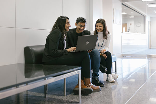 three students sit on a bench, one holds laptop for the others to see the screen