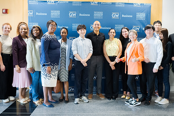 large, diverse group of business students take a group photo in front of GW Business step-and-repeat backdrop