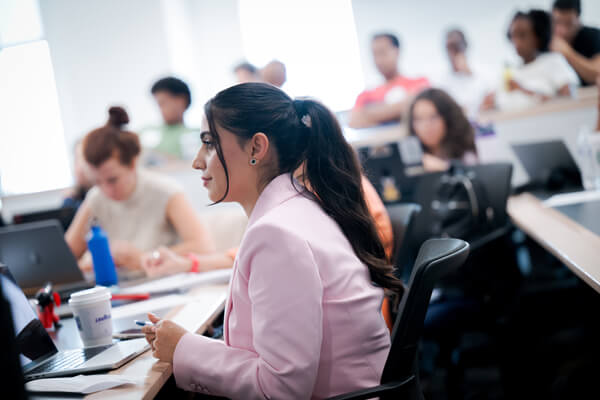 side shot of student wearing a light pink business suit jacket in class, other students blurred in background