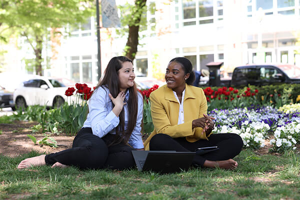 two students sit in the grass next to each other talking, flowers in the background
