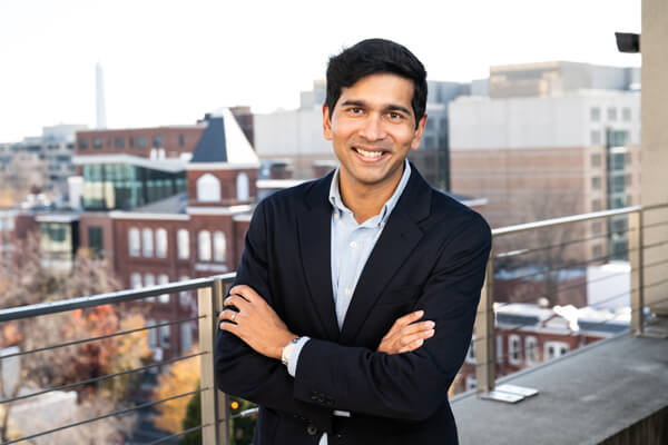 male student wearing a suit with arms crossed in front of him smiles directly at camera, campus buildings in the background