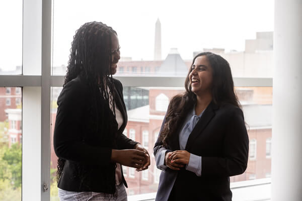 two students laugh in conversation, background is campus buildings through the big windows