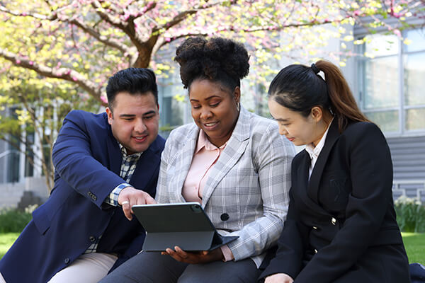three students sit on a bench outside next to each other, center student holds a laptop in her lap while the others look at the screen, one points to something