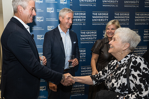 four professionals greet each other at event, two in the foreground shake hands, blue GW step-and-repeat in the background