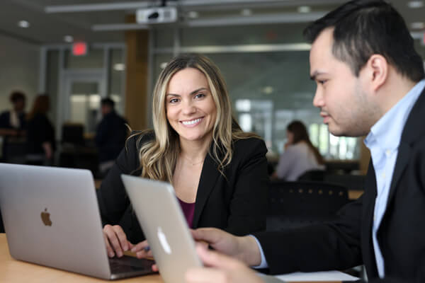 two students sit next to each other with laptops open in front of them, one smiles toward camera