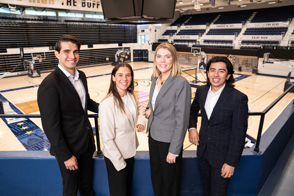 four students wearing business attire pose for a group photo with the GW basketball court in the background