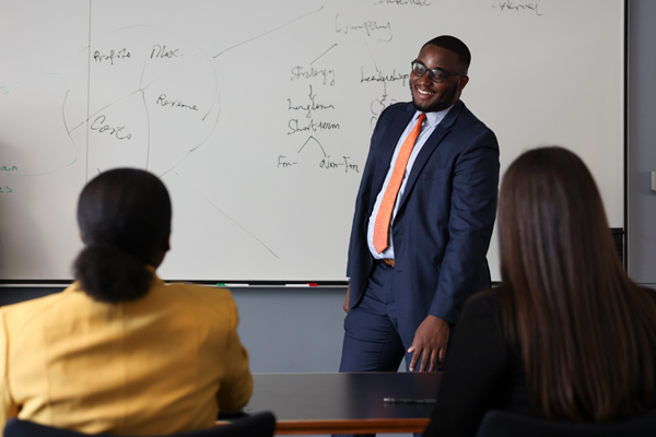 professor standing at front of class at whiteboard smiles, foreground includes the backs of two students
