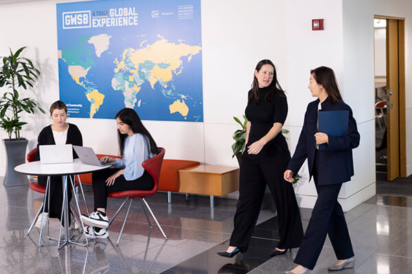 two professionals walk through the business school lobby, world map titled "GWSB Global Experiences" on the wall in background