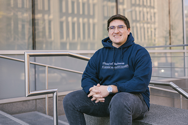 student wearing GW Business hoodie is seated on concrete wall outside, smiles to someone off camera