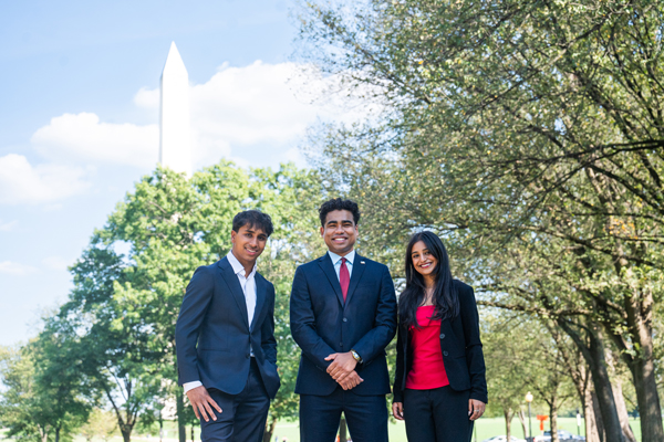 three international business students pose for a photo smiling at the camera, Washington Monument peaks above trees in background