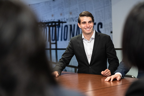 focus is on male student wearing black suit seated at table, the backs of two students heads are in the foreground of the photo