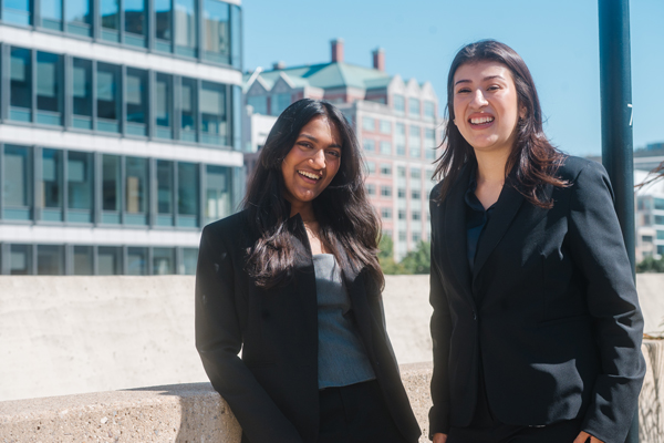 two female marketing students smile at camera while standing on outdoor rooftop patio, large buildings in the background