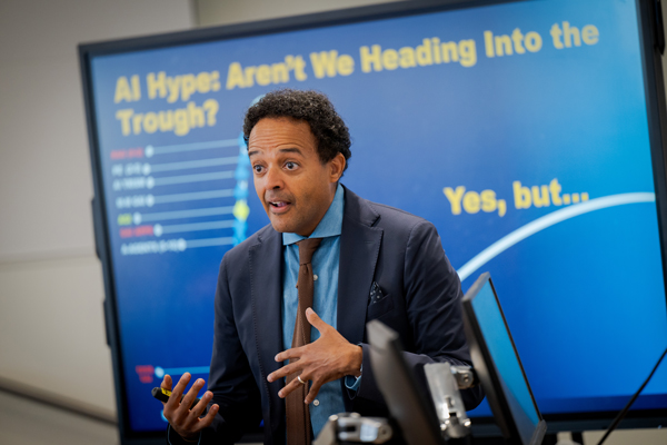 faculty member stands in front of class teaching, screen behind him reads "AI Hype: Aren't we heading into the trough? Yes, but..."