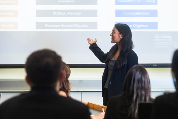 student stands in front of classroom giving presentation, turned and points to slide on digital screen behind her