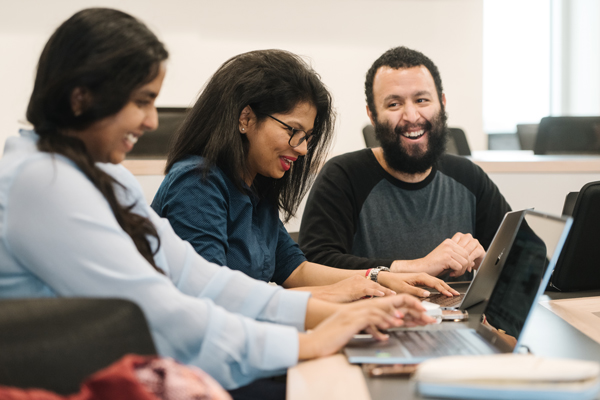 three students laugh together during class while working on laptops in front of them