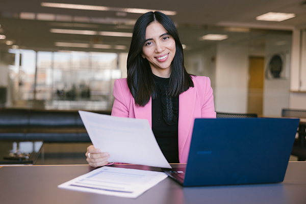 student wearing black top and pink suit jacket smiles at camera, holding a piece of paper in one hand, laptop open on her left