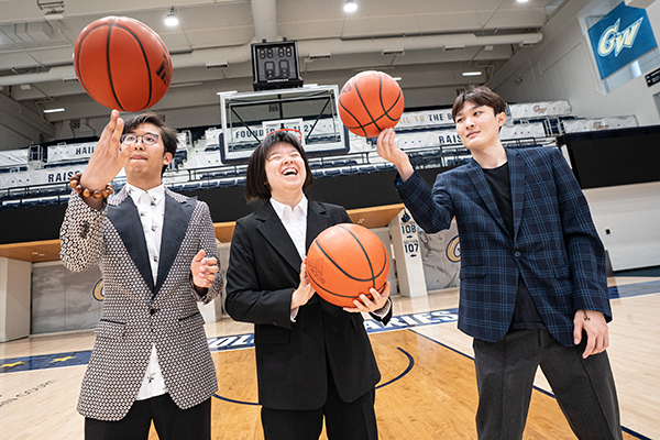 three students wearing suits balance basketballs on their fingers (one holding ball in hand) on the GW basketball court